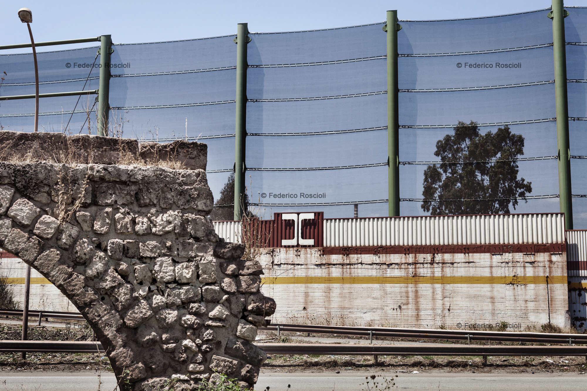 Taranto, Italy, August 1, 2013. Enclosure of Ilva factory in Taranto. The fence   over the wall was built in 2012 to protect the near neighborhood of Tamburi from the mineral dust coming from the implant. In foreground, the remains of the ancient Triglio aqueduct, belonging to Roman's eve. The Tamburi (drums) neighborhood is called so due to the sound made by the water dropping from the aqueduct.